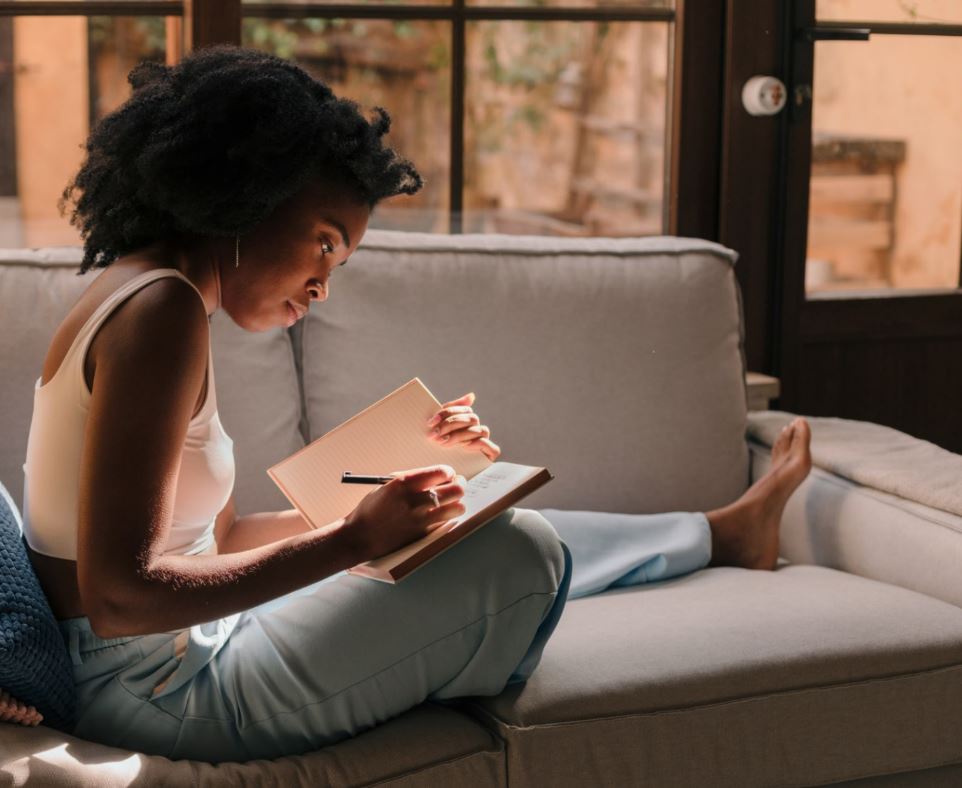 A young woman sitting on a couch in a comfy position writing in her notebook